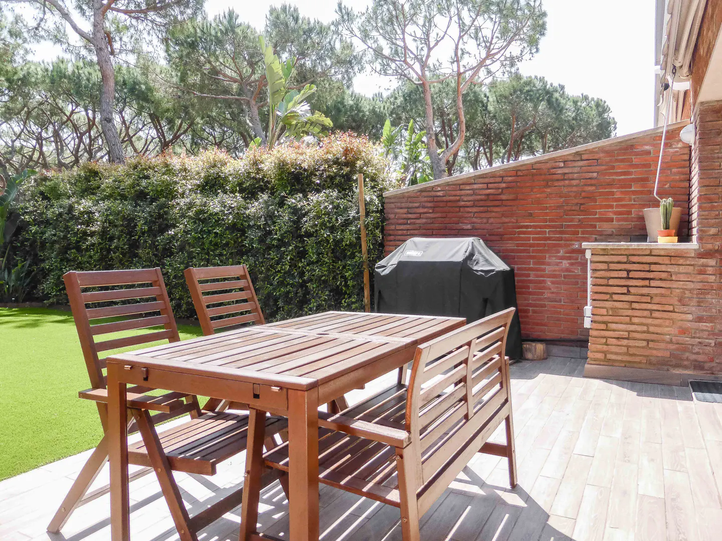 Outdoor patio with wooden table and chairs, a covered grill, and a brick wall with greenery in the background.