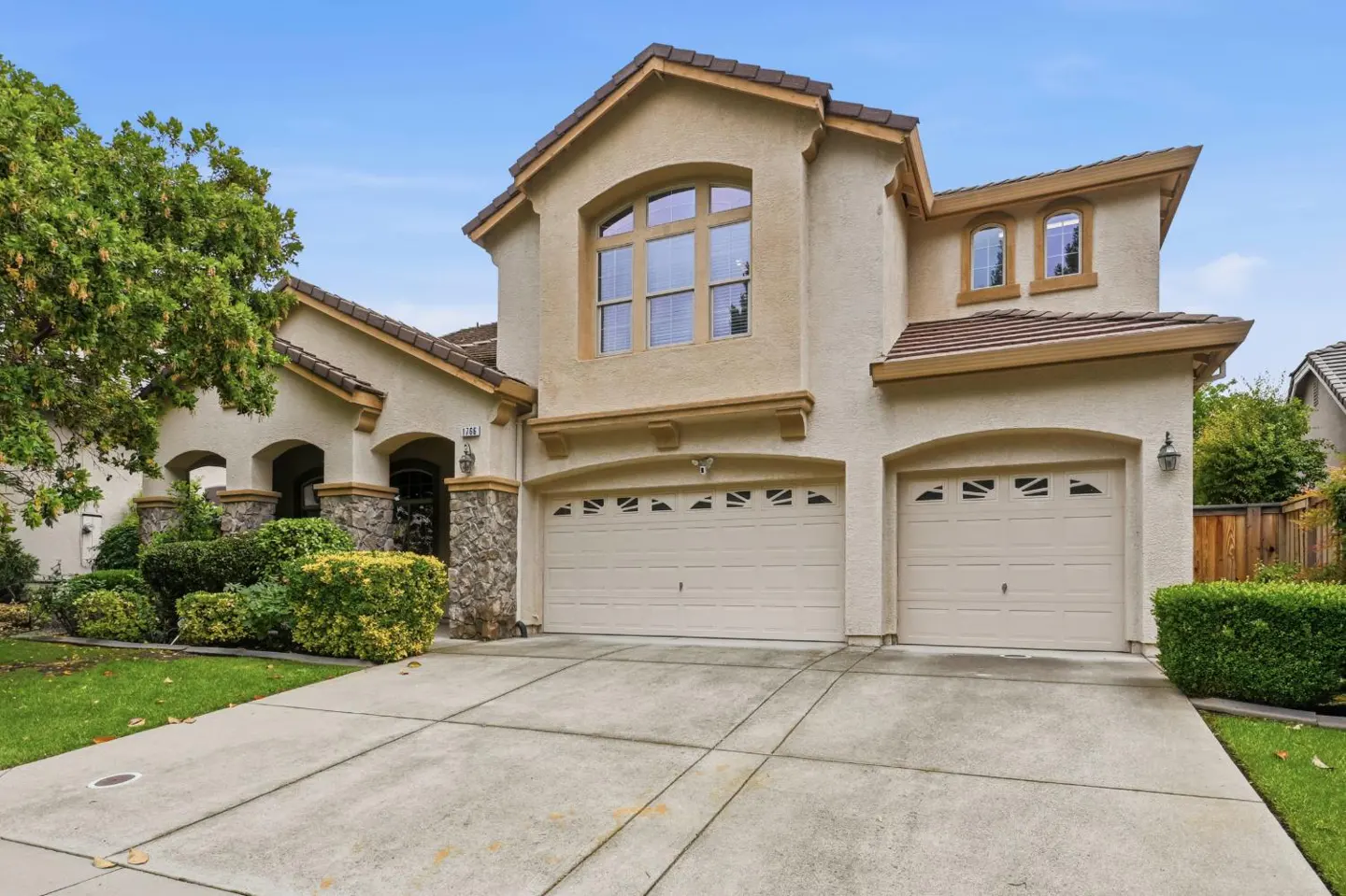 Two-story beige house with a brown roof and a two-car garage, green lawn, and blue sky.