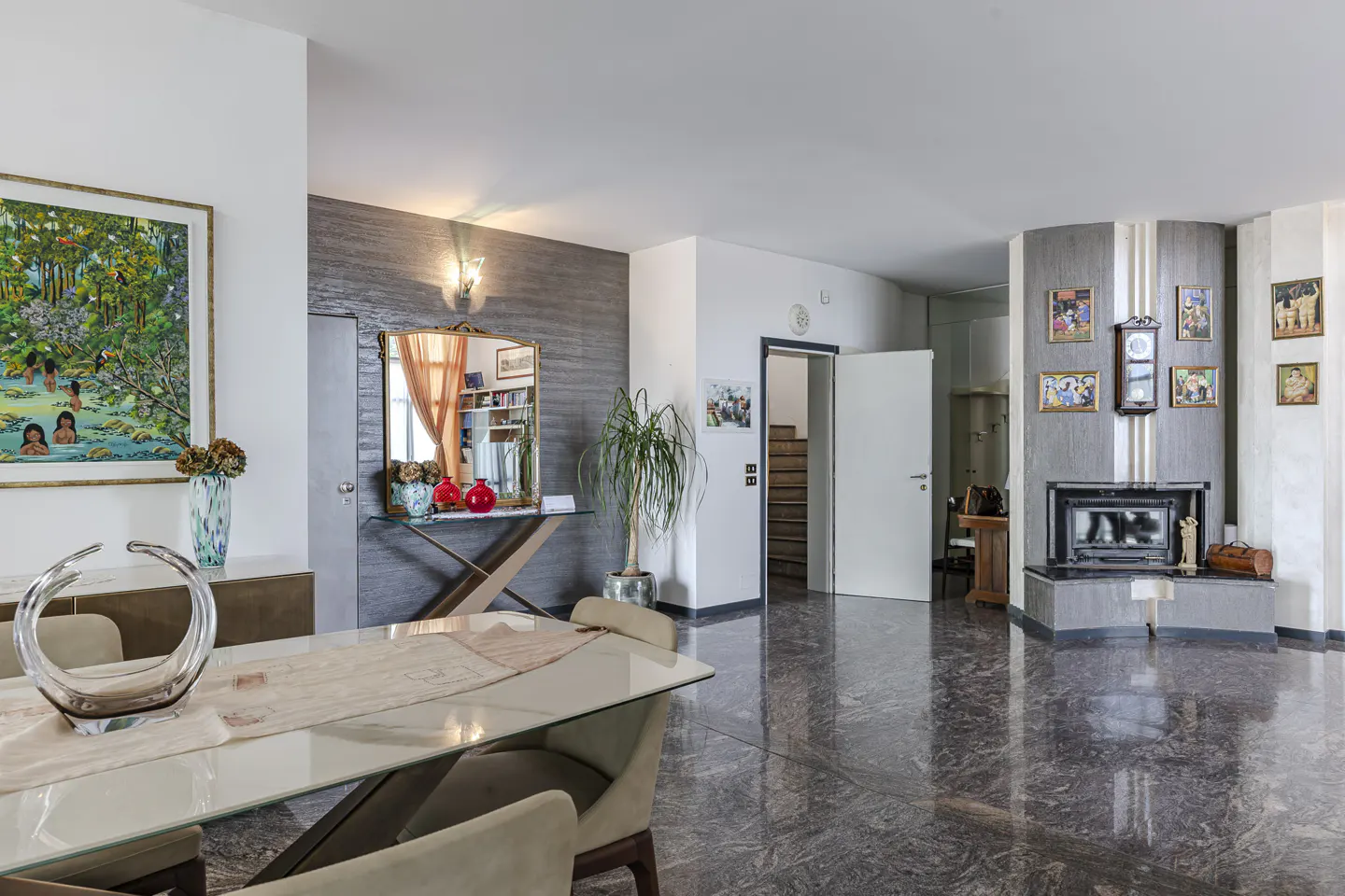 Modern dining room with glass table, marble floors, fireplace, and colorful artwork on gray and white walls.