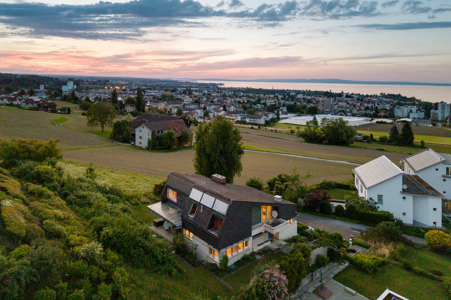 Aerial view of a modern house with a dark roof, green yard, and a city and lake in the background at sunset.