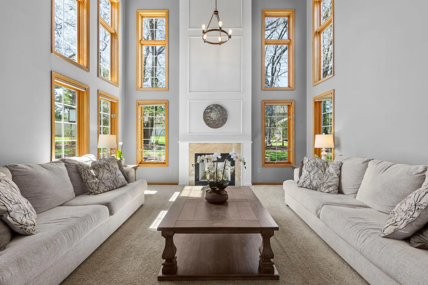 Bright living room with gray walls, light sofas, and a fireplace. Tall windows with wood trim frame the outdoor view.