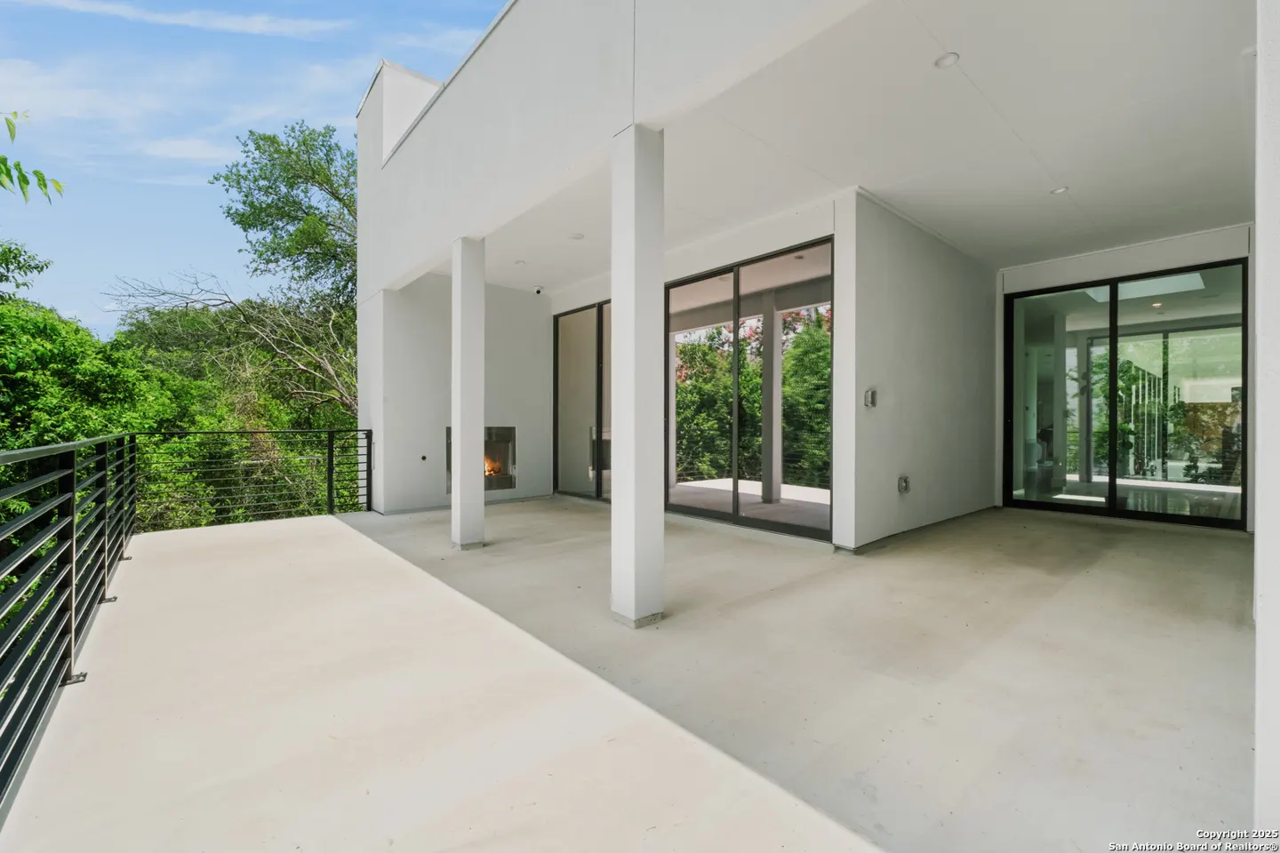 Wide shot of a modern white patio with black railings, columns, and sliding glass doors overlooking lush green trees.