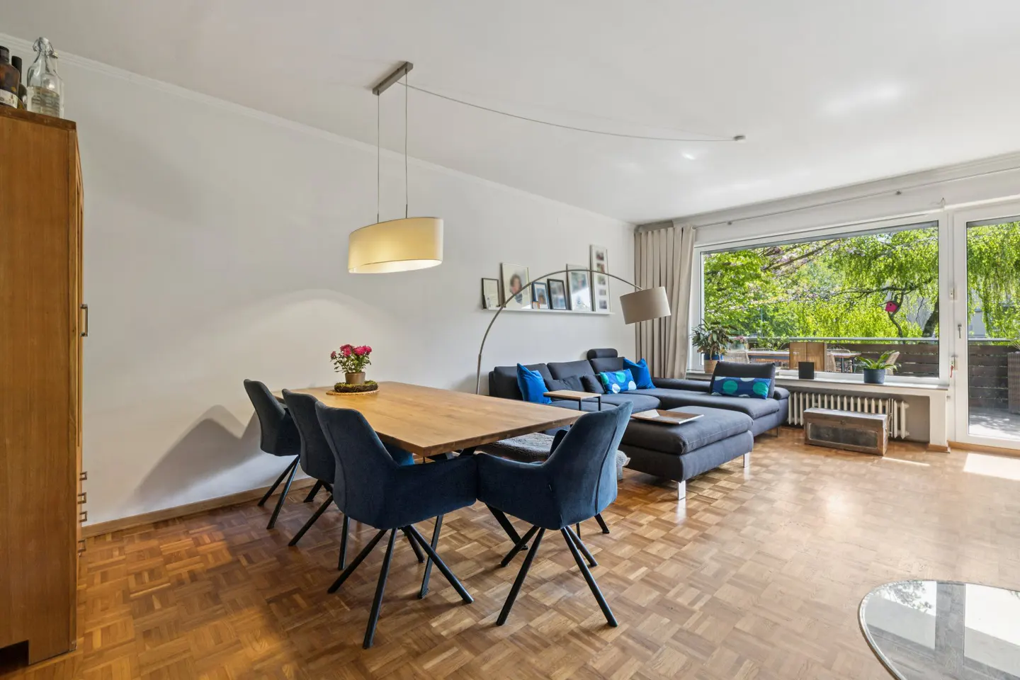 Bright living room with wood floors, a dining table with blue chairs, and a gray sectional sofa near a large window.