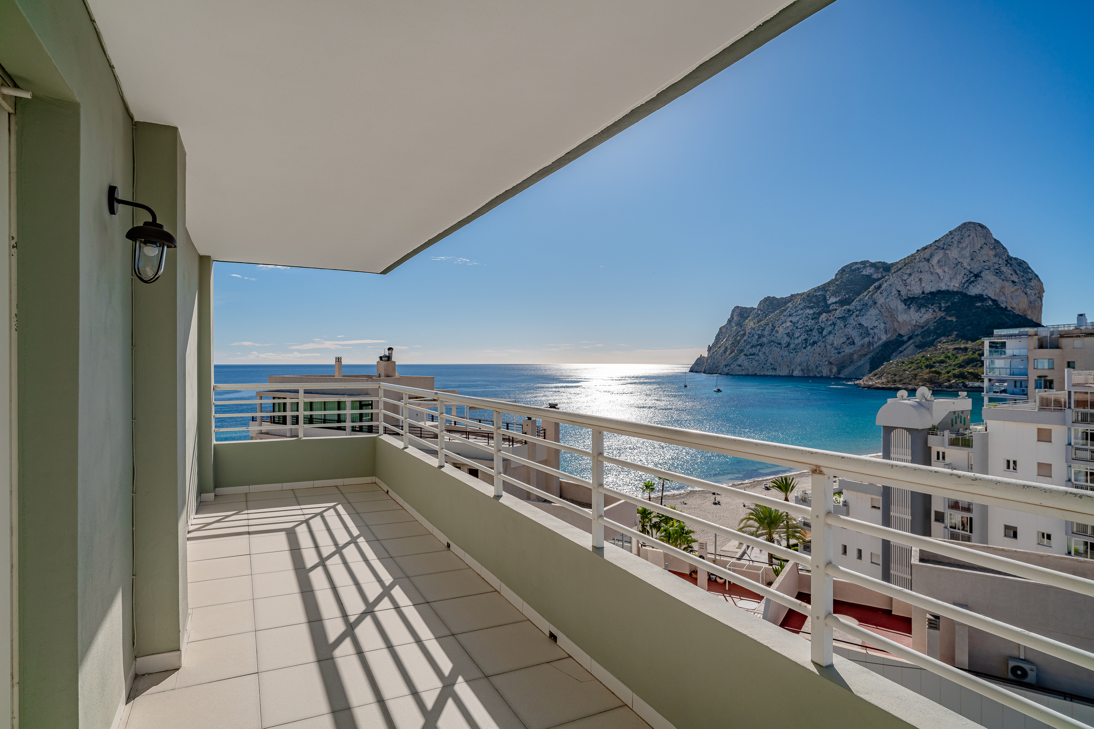 Balcony with white railings overlooking a sunny Mediterranean beach, blue sea, and a large rocky mountain under a clear sky. [AI-generated text]