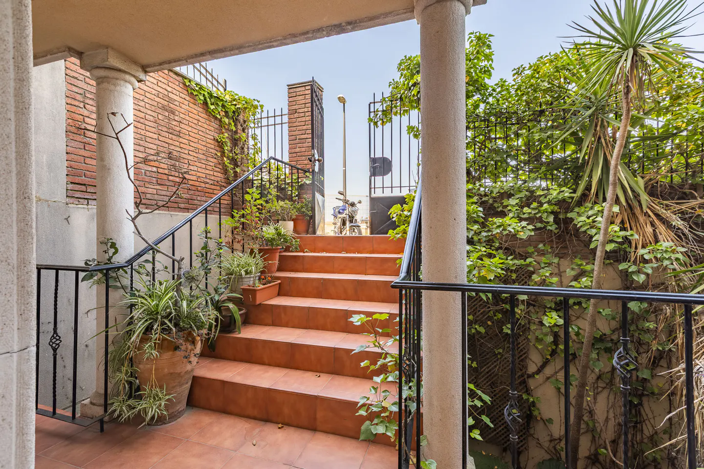 Outdoor staircase with terracotta tiles, black railings, and potted plants leading to an open gate with greenery.