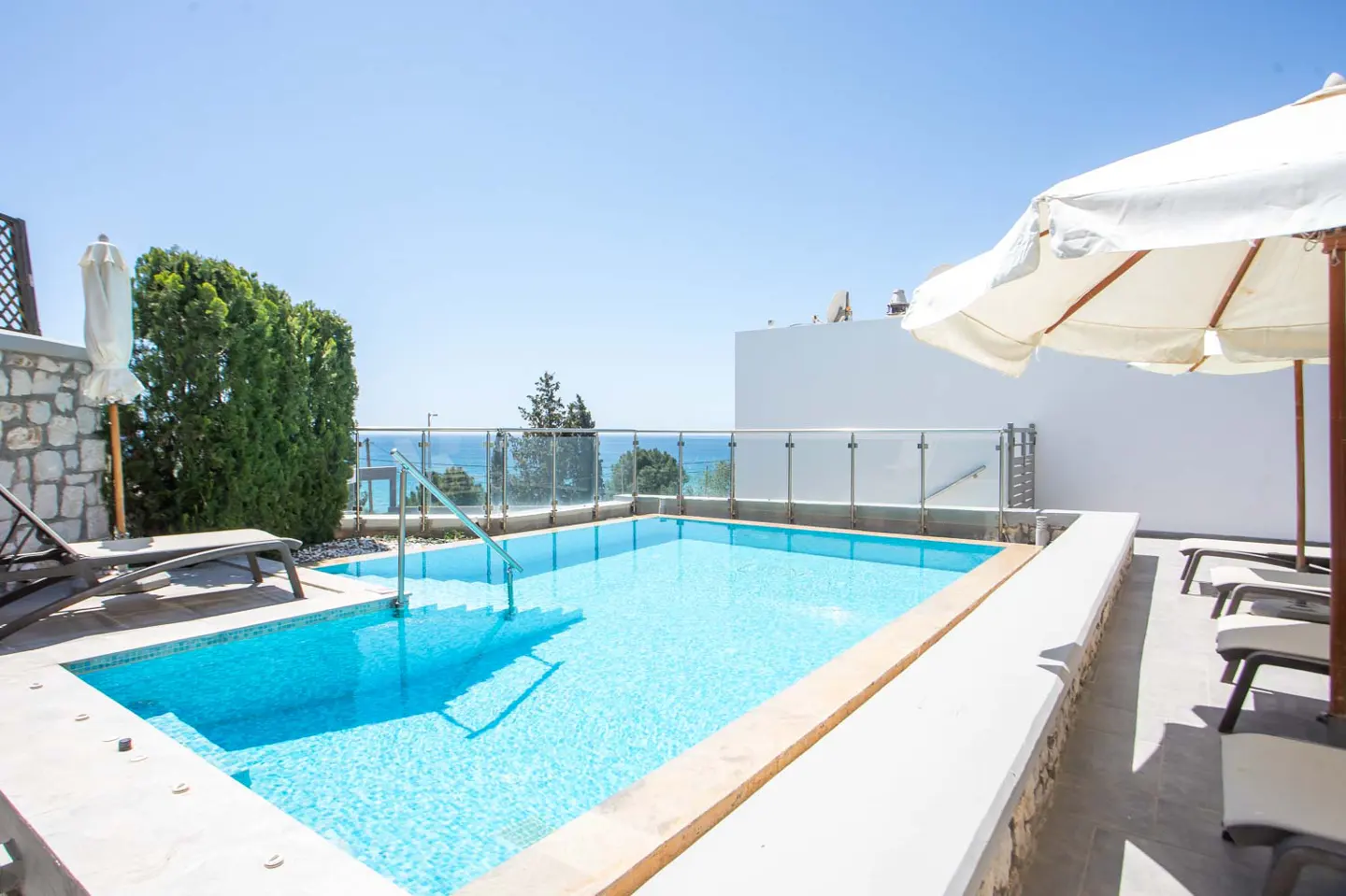 Bright blue tiled pool with steps, surrounded by white walls and lounge chairs, with ocean view and sun umbrellas.