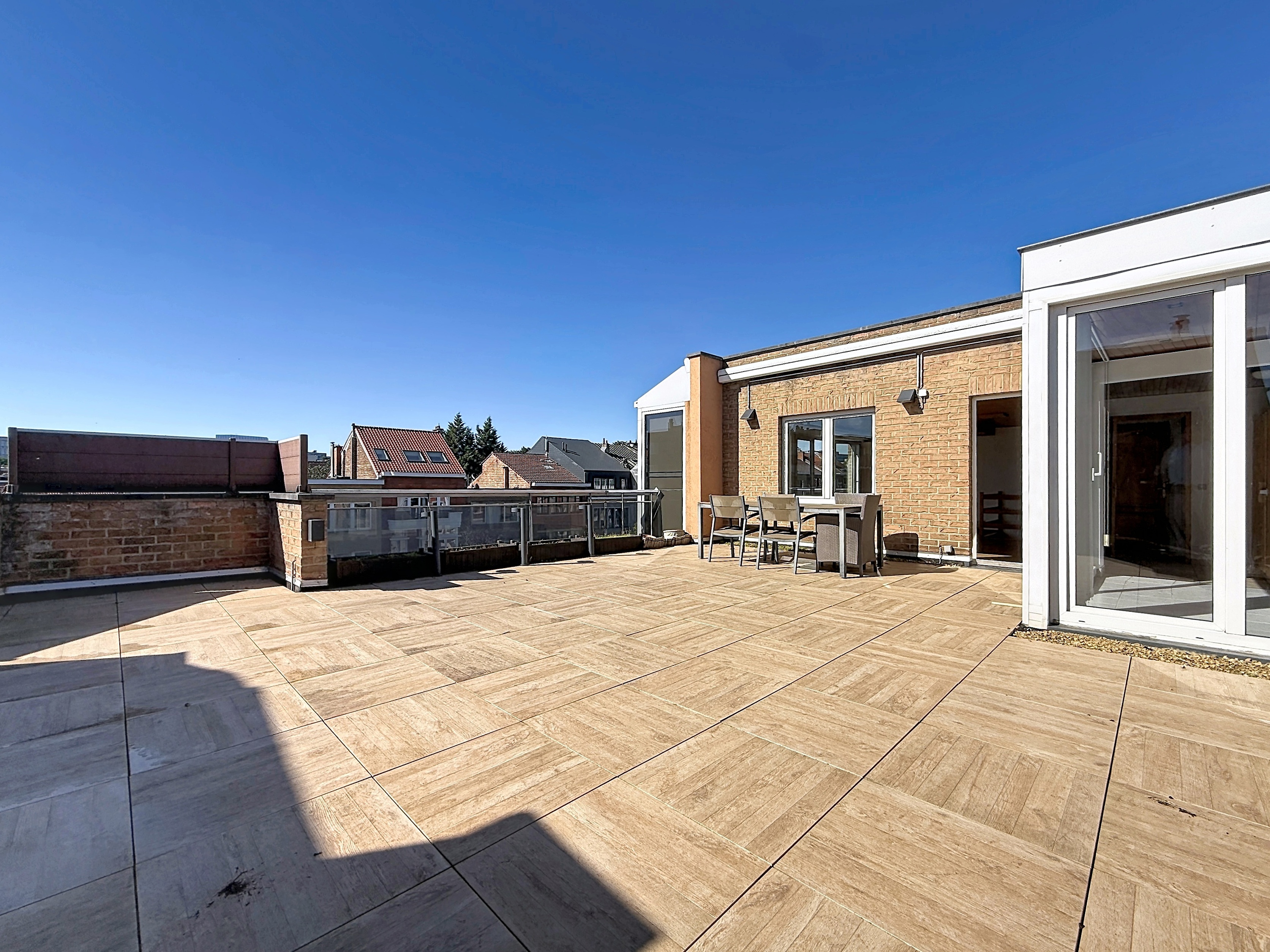 Spacious rooftop terrace with light wood-look tiles, outdoor dining set, brick walls, and clear blue sky. [AI-generated text]