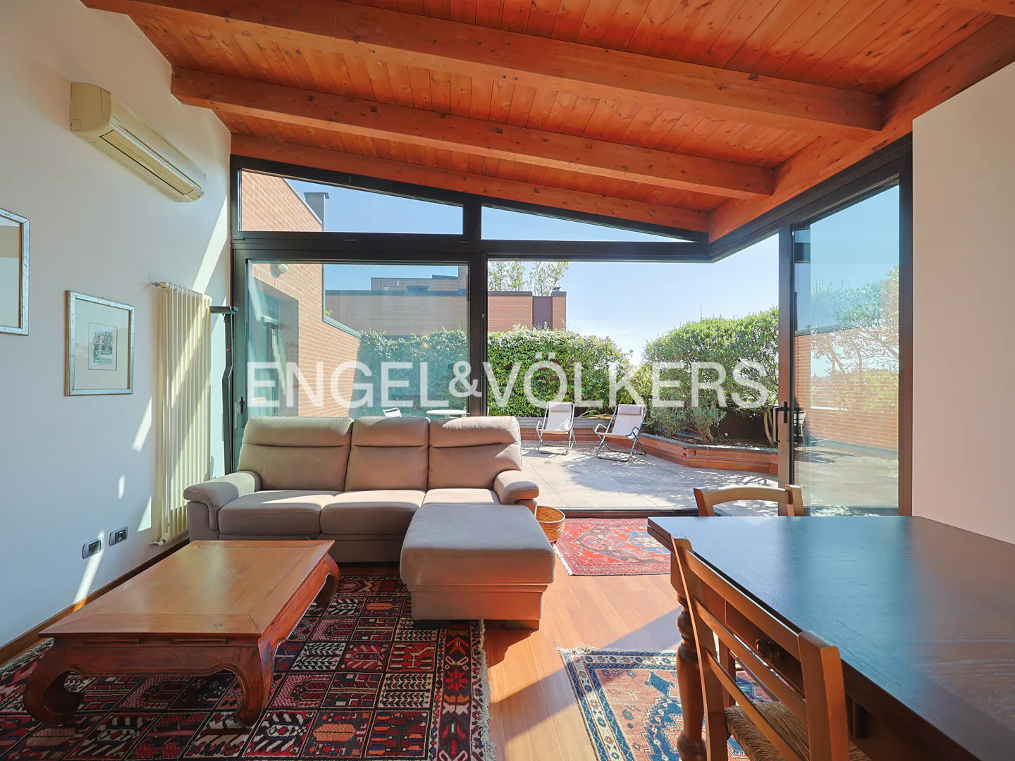 Living room with a beige sectional sofa, wooden coffee table, and patterned rugs under a vaulted wood-beam ceiling.