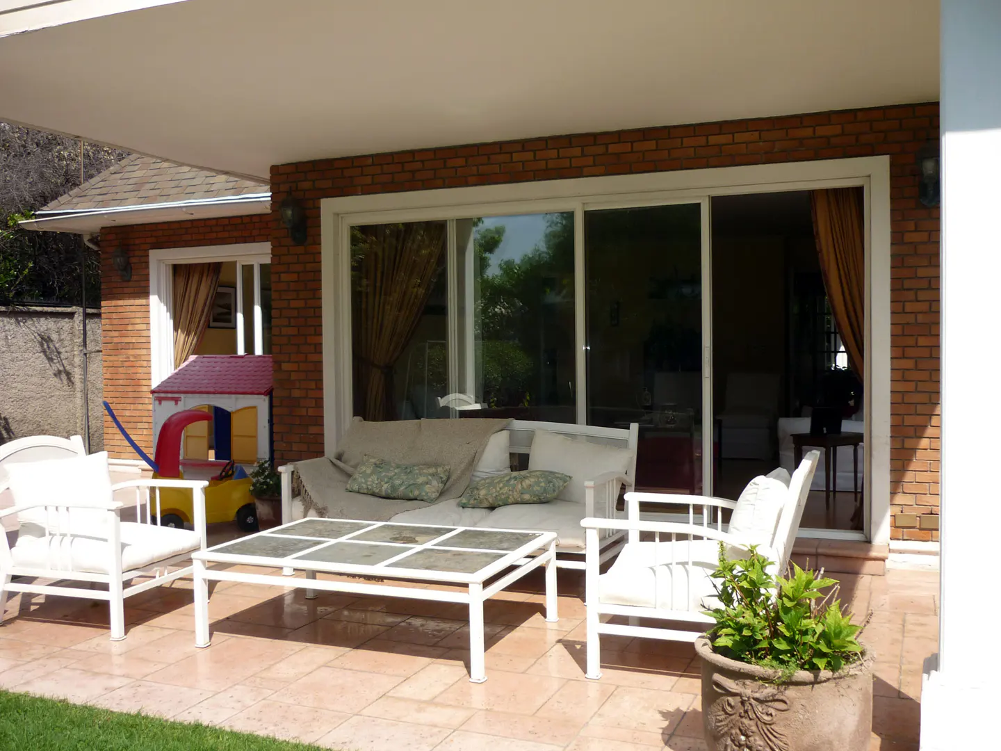 Covered patio with white furniture, including a sofa, chairs, and a coffee table. A red brick wall and a child's playhouse are in the background.