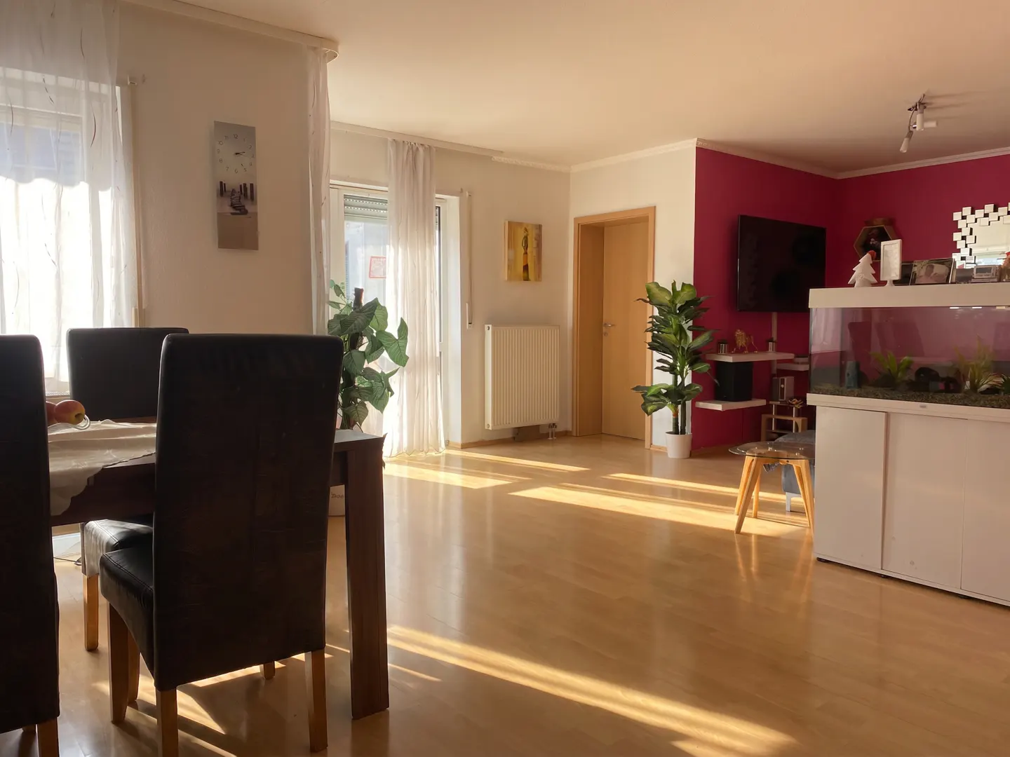 Bright, open-concept living and dining area with wood floors. Dining table with brown chairs in the foreground, aquarium and TV in the background.