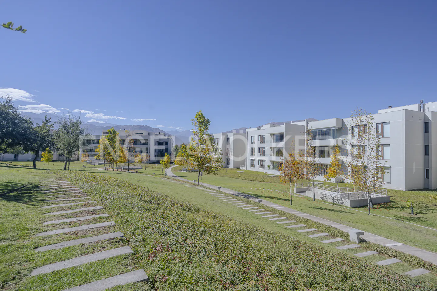 Exterior view of modern white apartment buildings with green lawns and stone pathways under a clear blue sky.
