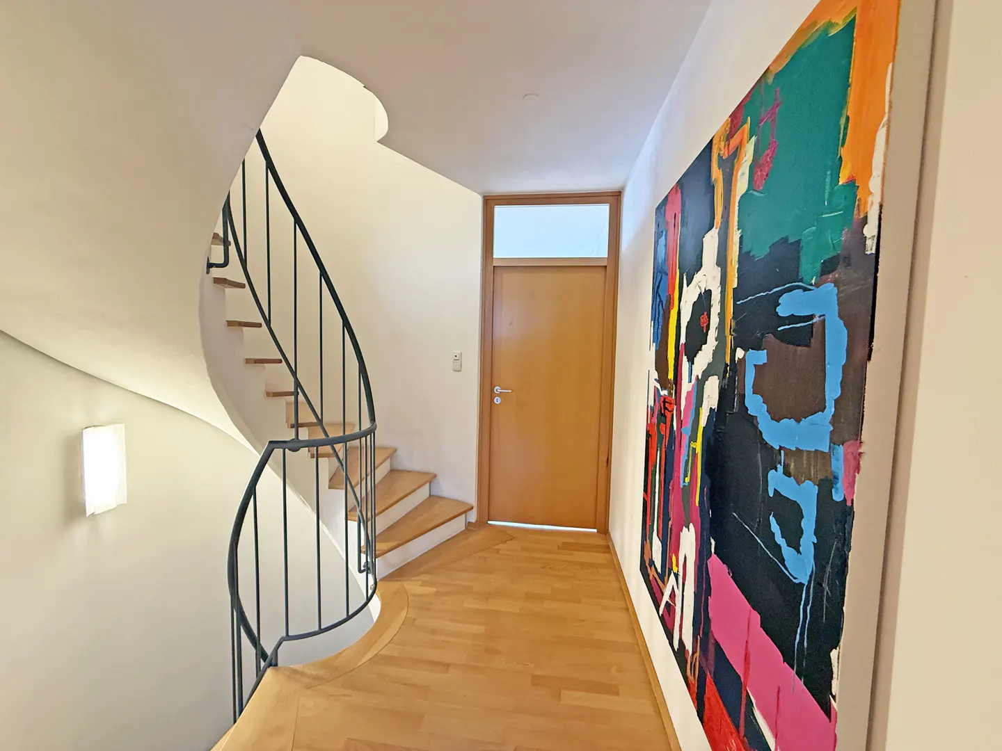 Hallway with a spiral staircase, wood floors, a door, and a large colorful abstract painting.