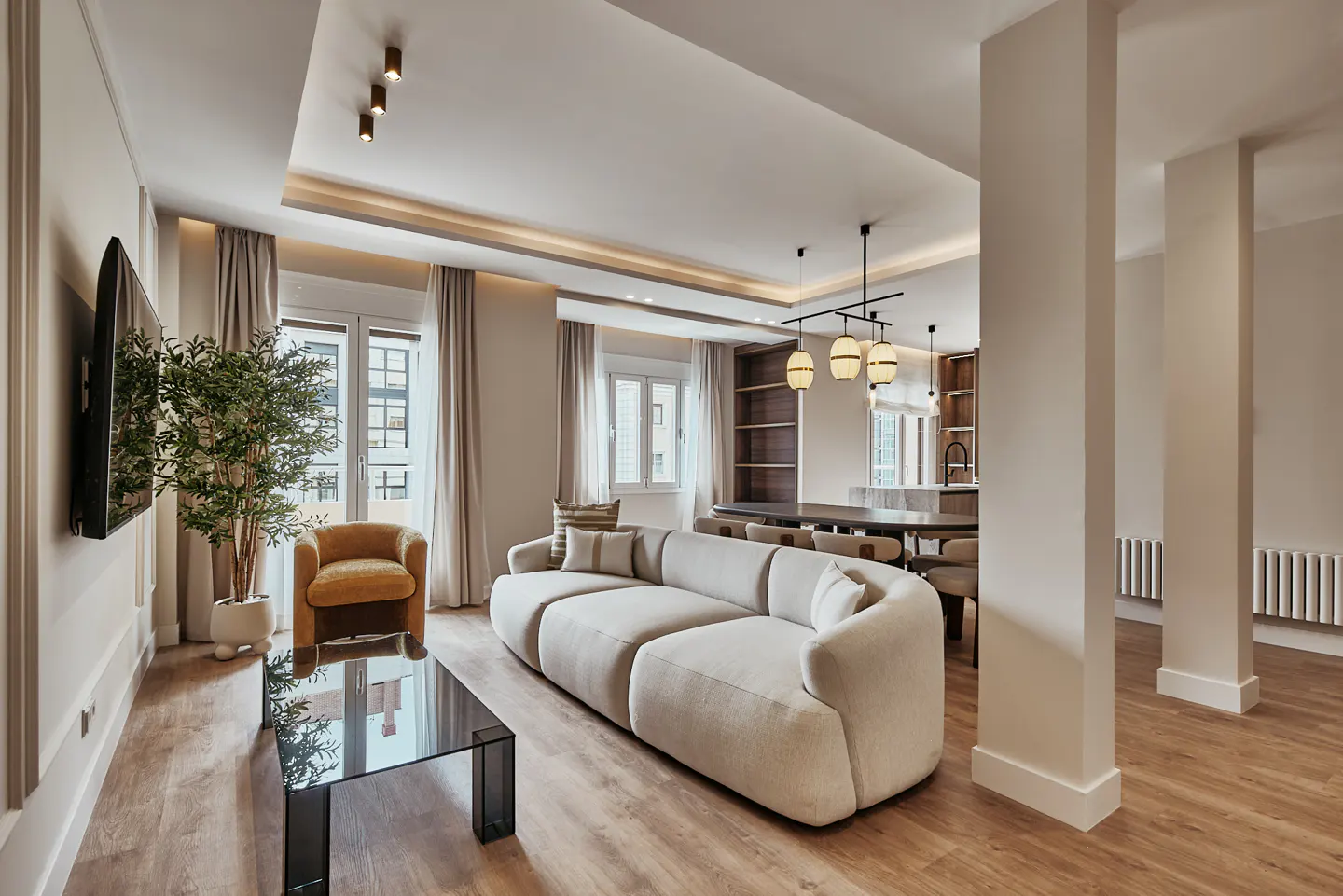 Modern open-plan living room with a beige curved sofa, glass coffee table, mustard armchair, and dining area in the back.