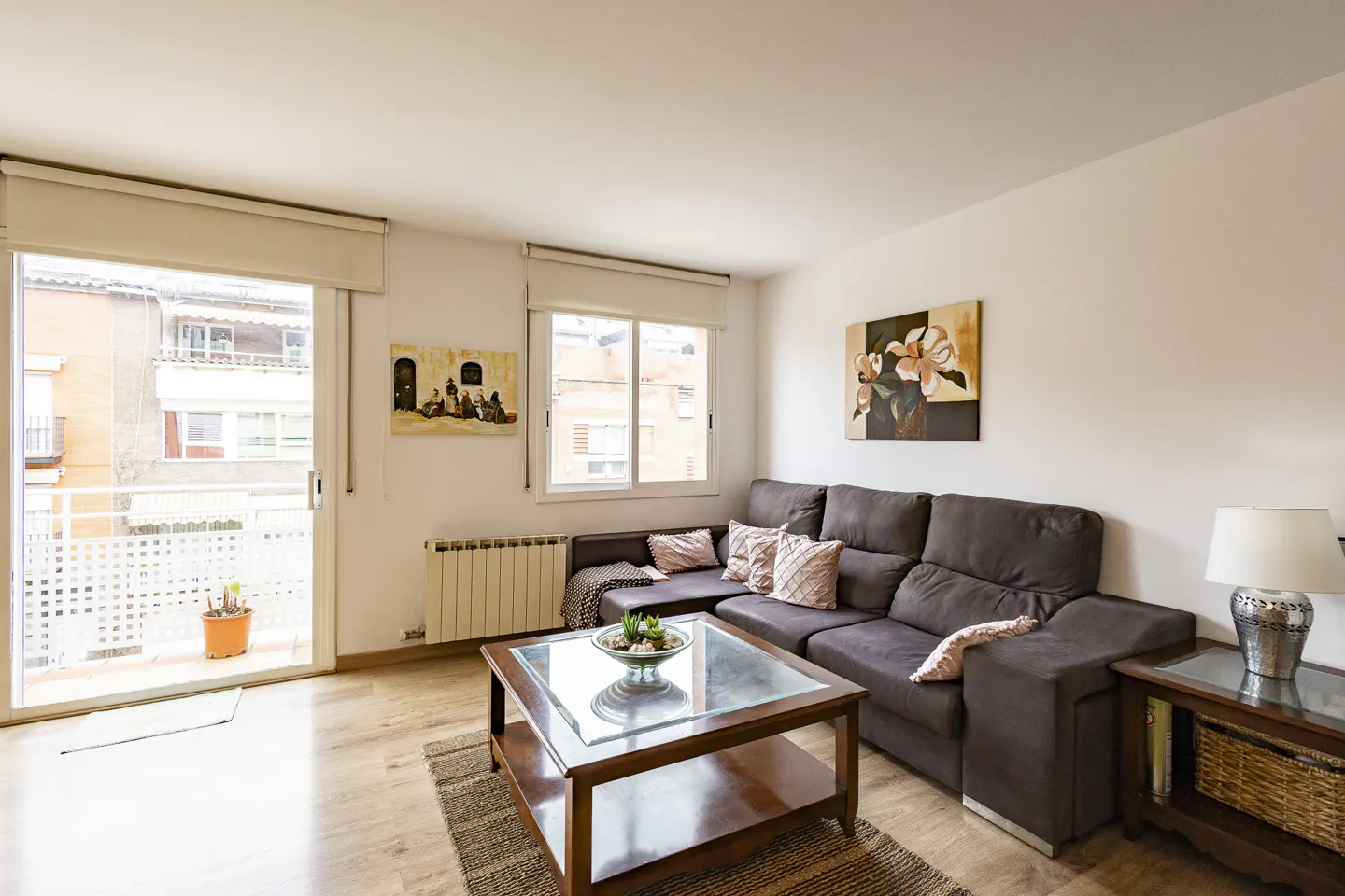 Bright living room with a brown sectional sofa, glass-topped coffee table, and balcony access. Paintings adorn the white walls.