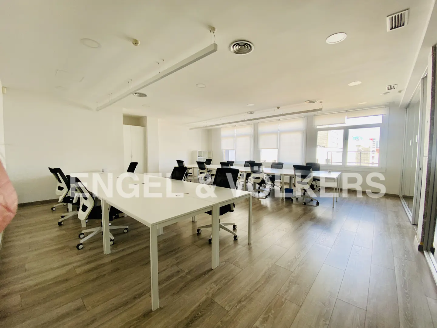 Bright office space with white walls, wood-look floors, and rows of white desks with black chairs. Windows provide natural light.