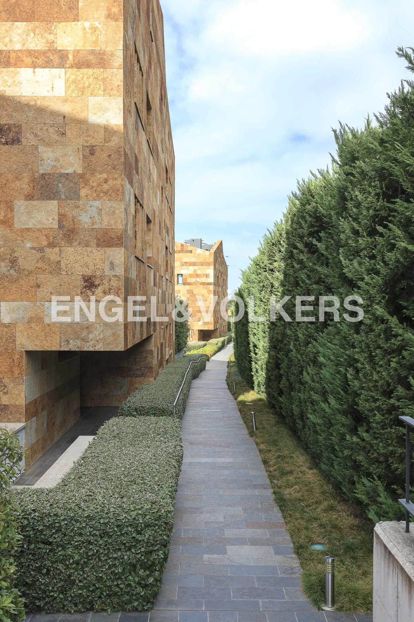 Stone walkway between modern buildings and tall hedges under a blue sky. Engel & Volkers logo visible on the building.