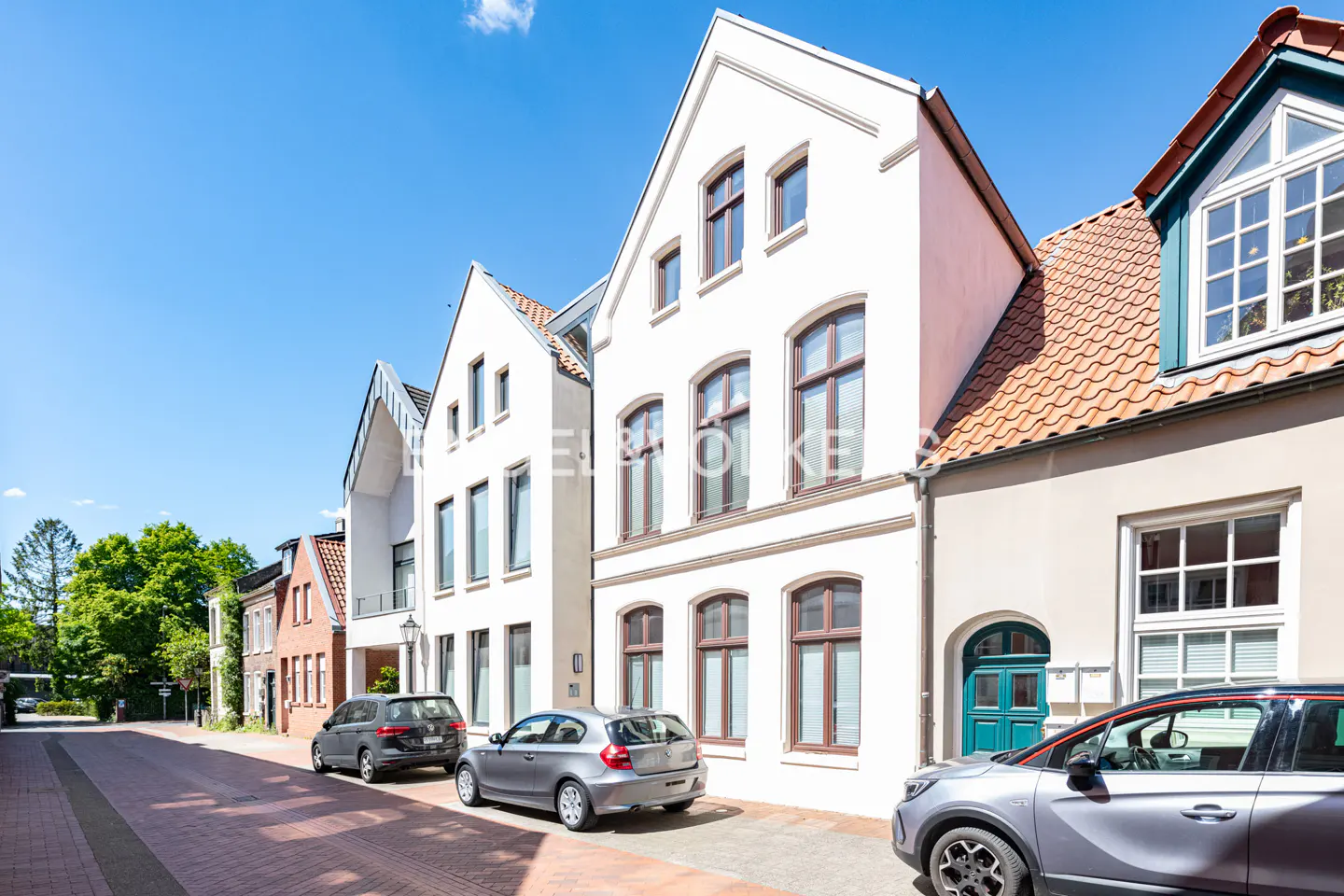 Street view of white, multi-story townhouses with parked cars on a brick road under a clear blue sky.
