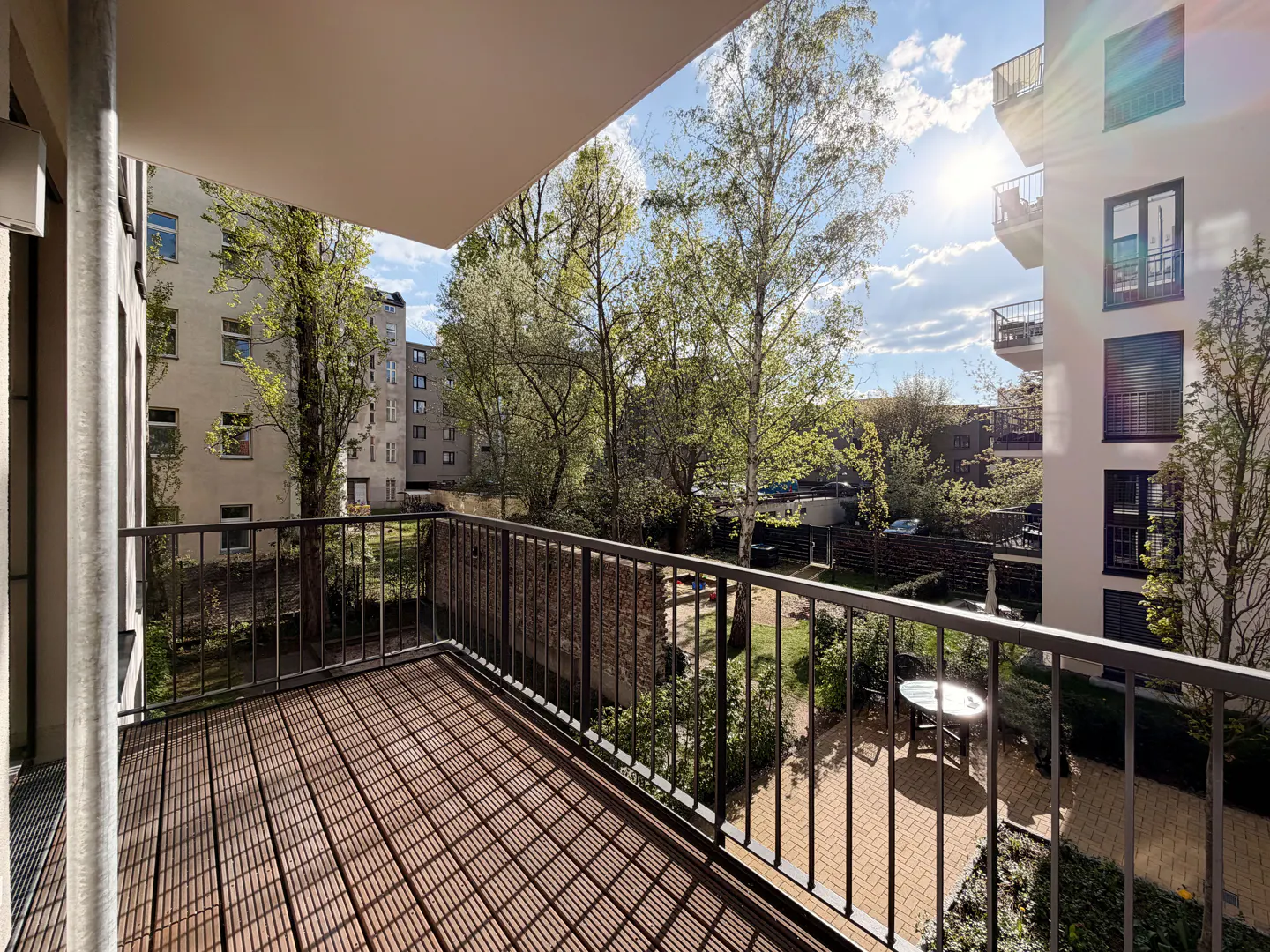 Balcony with brown wood floor and black metal railing overlooking a courtyard with trees, grass, and a table. Buildings are visible in the background.