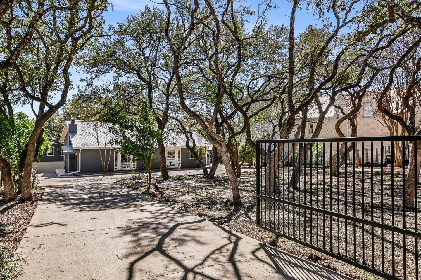 Gated driveway leads to a gray house with a metal roof, framed by trees casting shadows on the concrete.