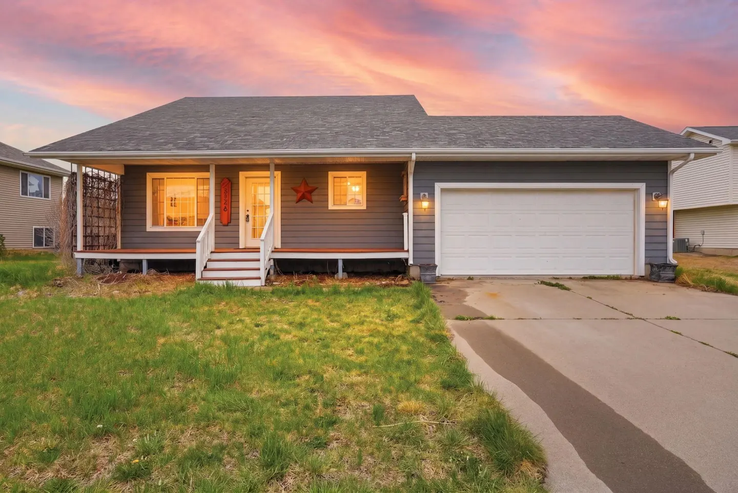 A gray single-story house with a white garage door, porch, and a red star decoration. The sky is pink and purple.