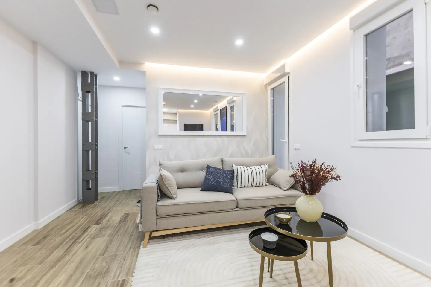 Living room with beige sofa, pillows, and two black nesting tables with a vase of flowers on a white rug.