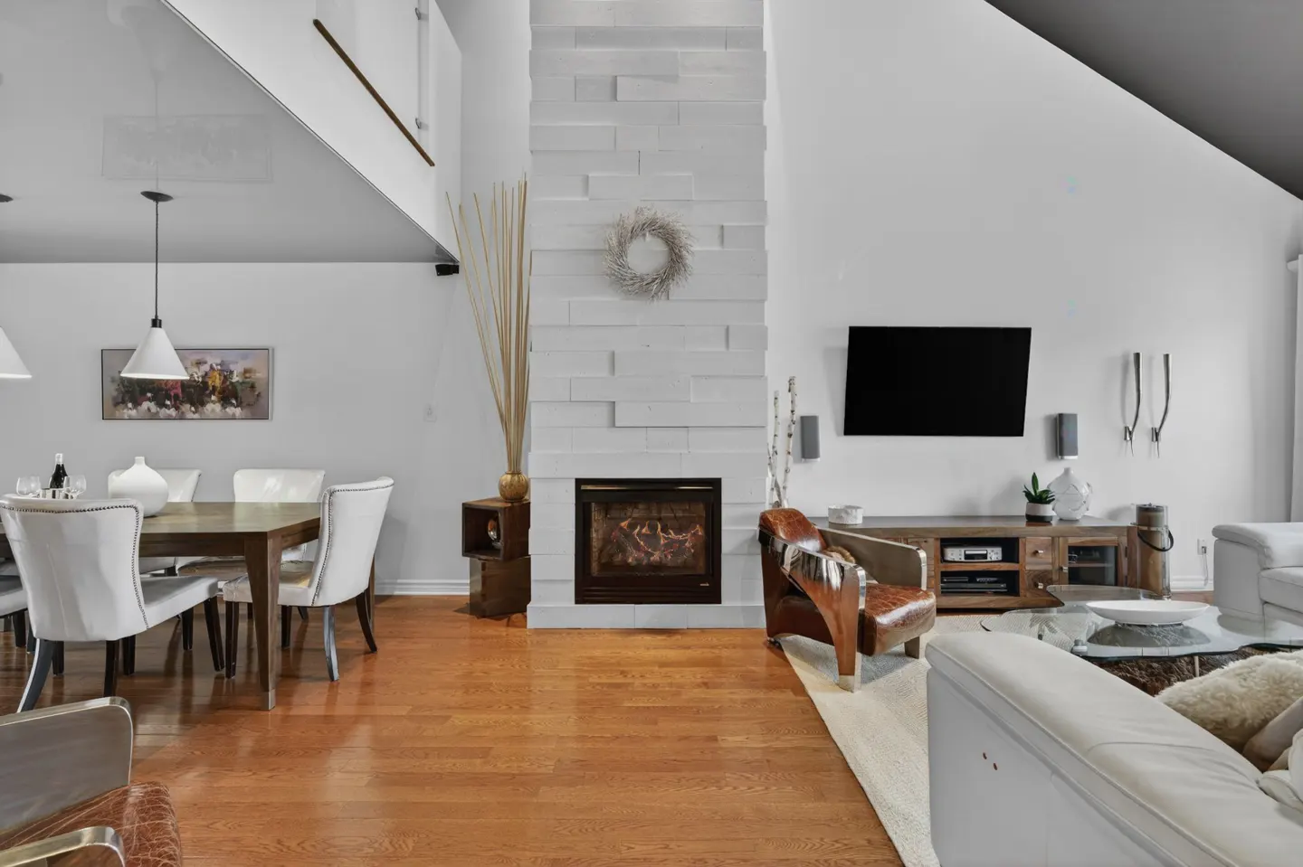 Bright living room with hardwood floors, white walls, and a modern fireplace. A dining table with white chairs is on the left, and a TV and leather chair are on the right.