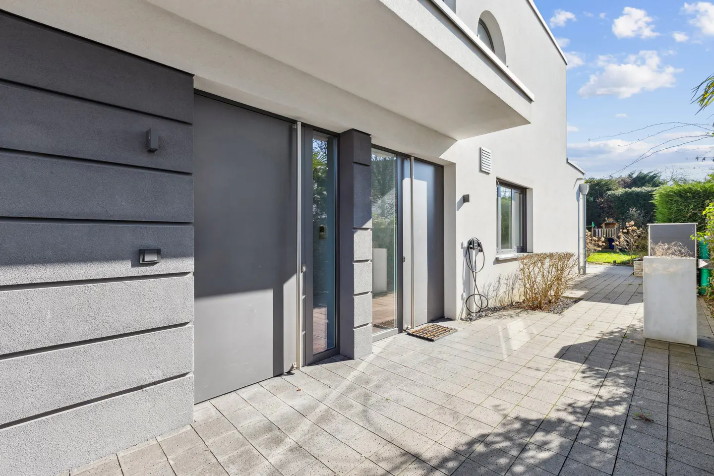Modern home exterior with gray siding, glass doors, and a paved patio. A charging cable is visible near a window.