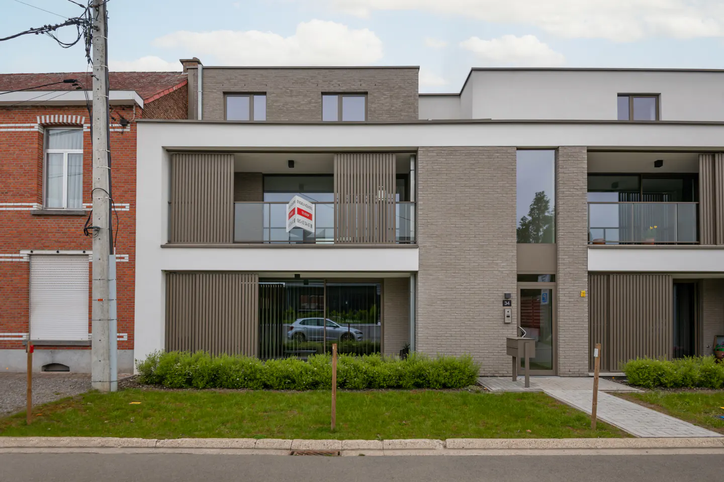 Modern townhouse with gray brick and white accents. A "For Sale" sign hangs on the balcony. Green lawn and walkway in front.