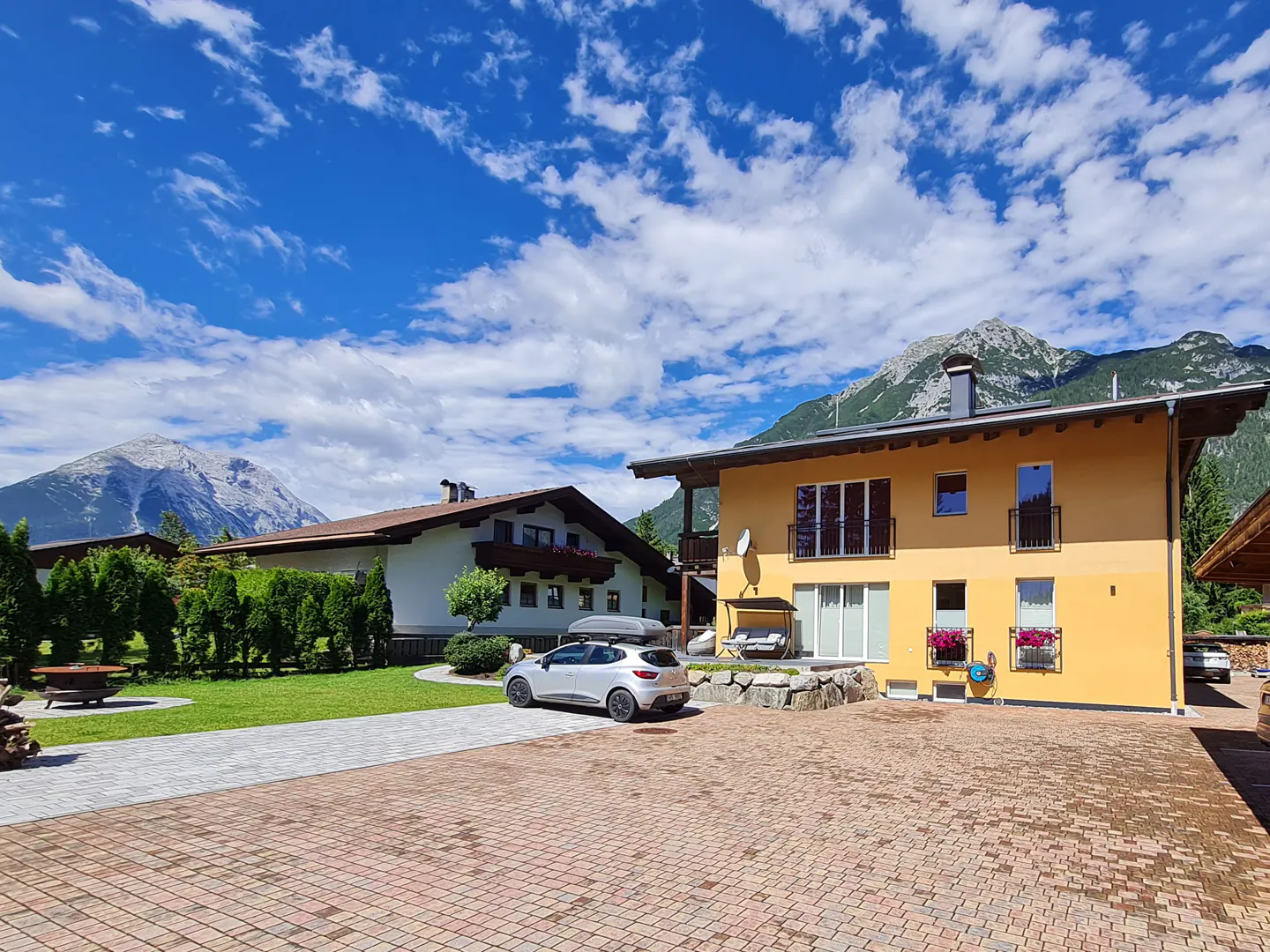 Two-story yellow house with black balconies, a car parked in front, and mountains in the background under a blue sky.