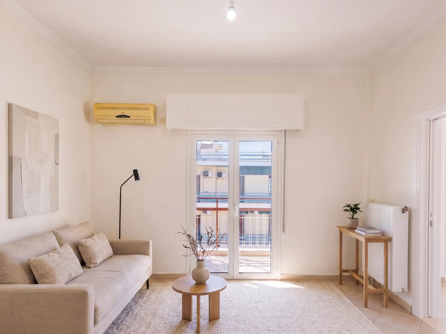 Bright living room with beige sofa, round wood table, and white walls. Balcony doors offer city views. Artwork and a floor lamp add detail.
