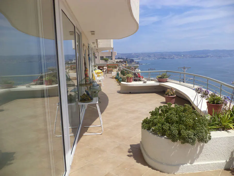 A sunny balcony with potted plants overlooks a blue ocean and distant cityscape. Sliding glass doors lead inside.