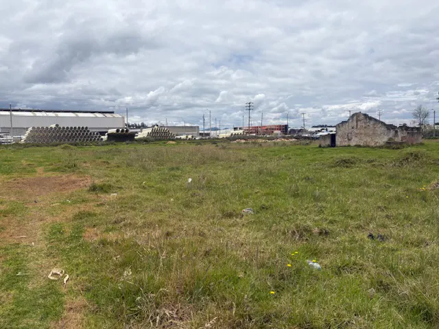 Vacant grassy lot with a dilapidated stone building and industrial buildings under a cloudy sky.