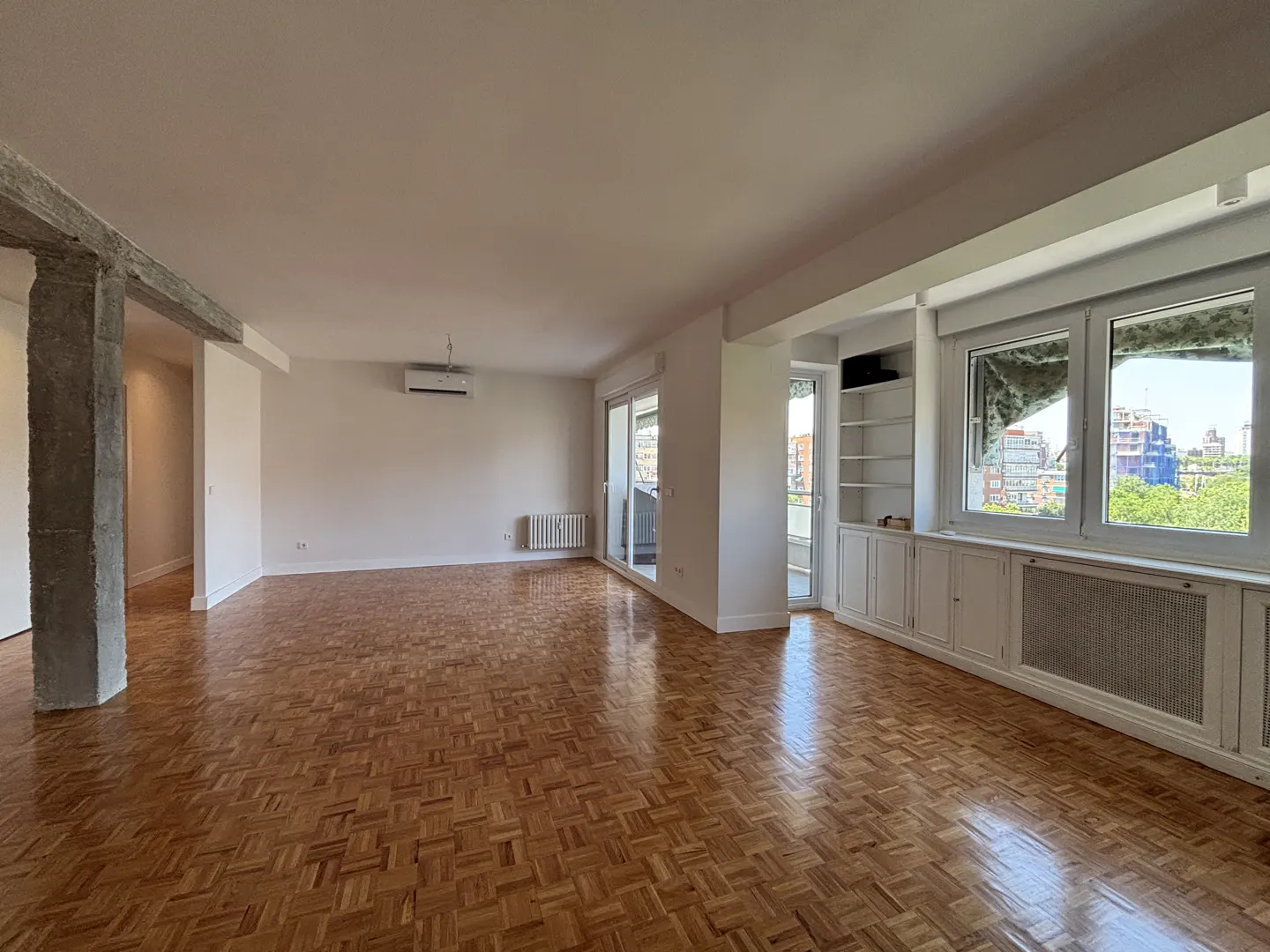 Spacious, empty living room with polished parquet wood floors, white walls, built-in shelving, and large windows.