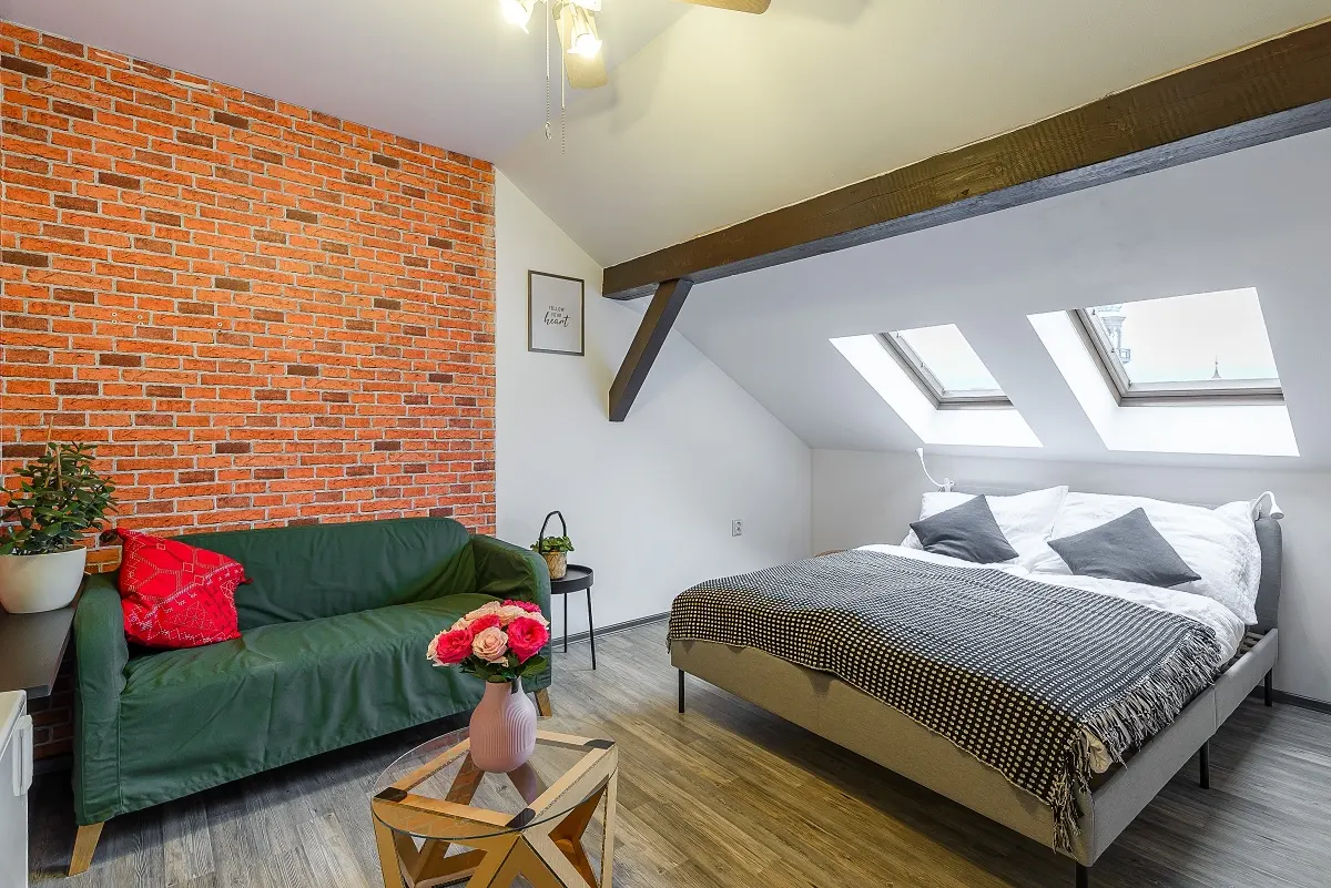 Attic bedroom with a red brick wall, green sofa, double bed with a patterned throw, and two skylights under wooden beams.