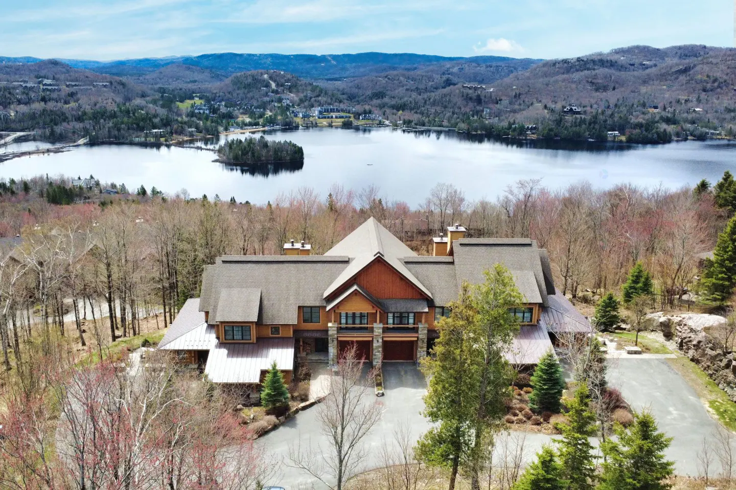 Aerial view of a large, multi-story brown wood and stone lodge nestled in a forest overlooking a scenic lake and mountains.