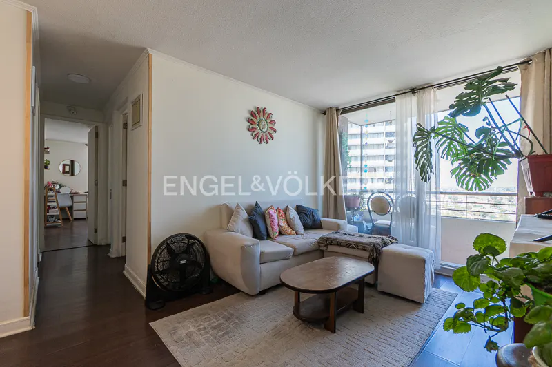 Living room with a beige sectional sofa, oval coffee table, and a black fan. Balcony with plants and a view of a city.