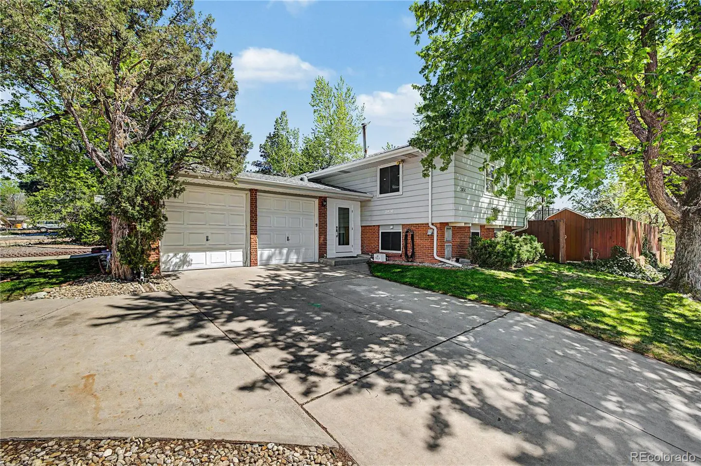 Two-story house with white siding, red brick foundation, and a two-car garage. Green trees frame the house.