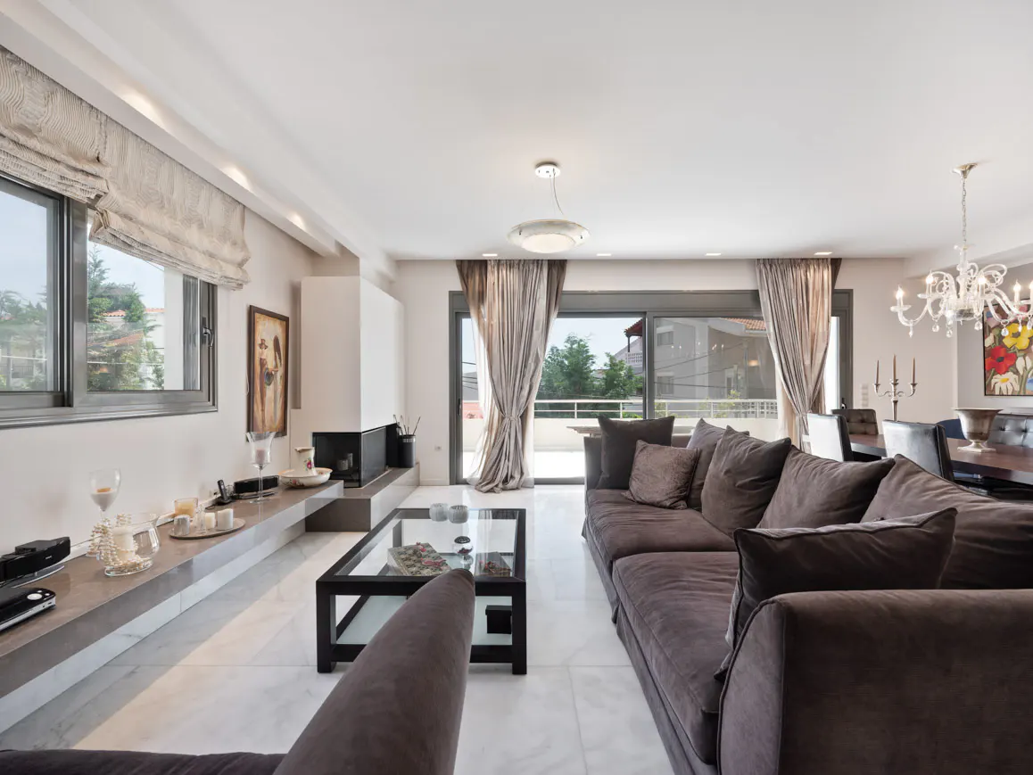 Bright living room with a large brown sofa, glass coffee table, fireplace, and marble floors. Windows and a balcony offer outdoor views.