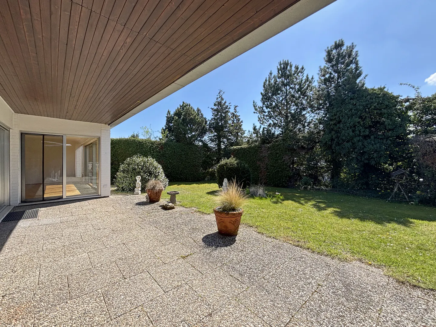 Patio view of a home with a brown wood ceiling, sliding glass doors, stone pavers, and a green lawn with trees.