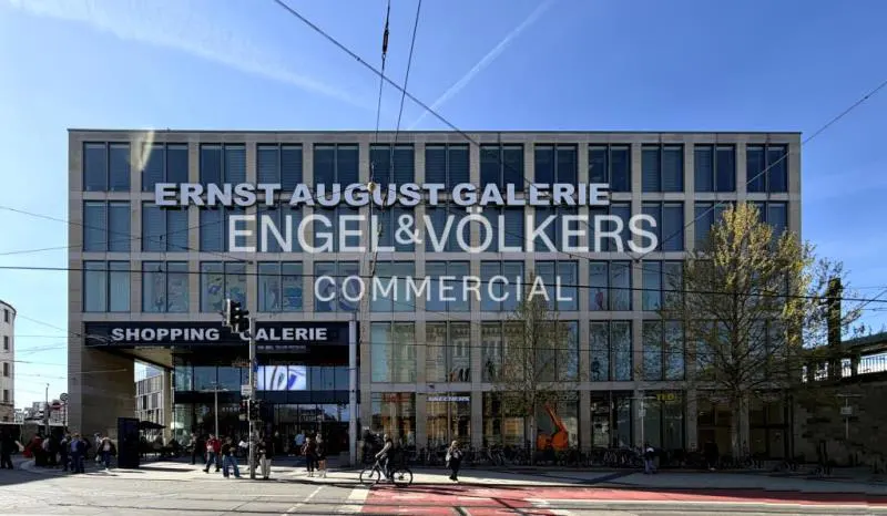 Exterior view of Ernst August Galerie, a commercial building with Engel & Völkers signage, on a sunny day. People walk and bike on the street.