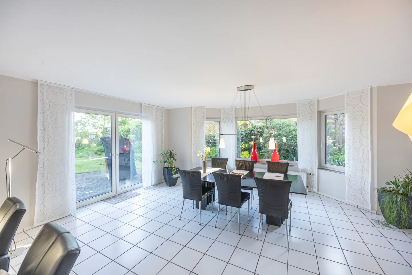 Bright dining room with white tile floor, black table and chairs, and large windows overlooking a green garden.
