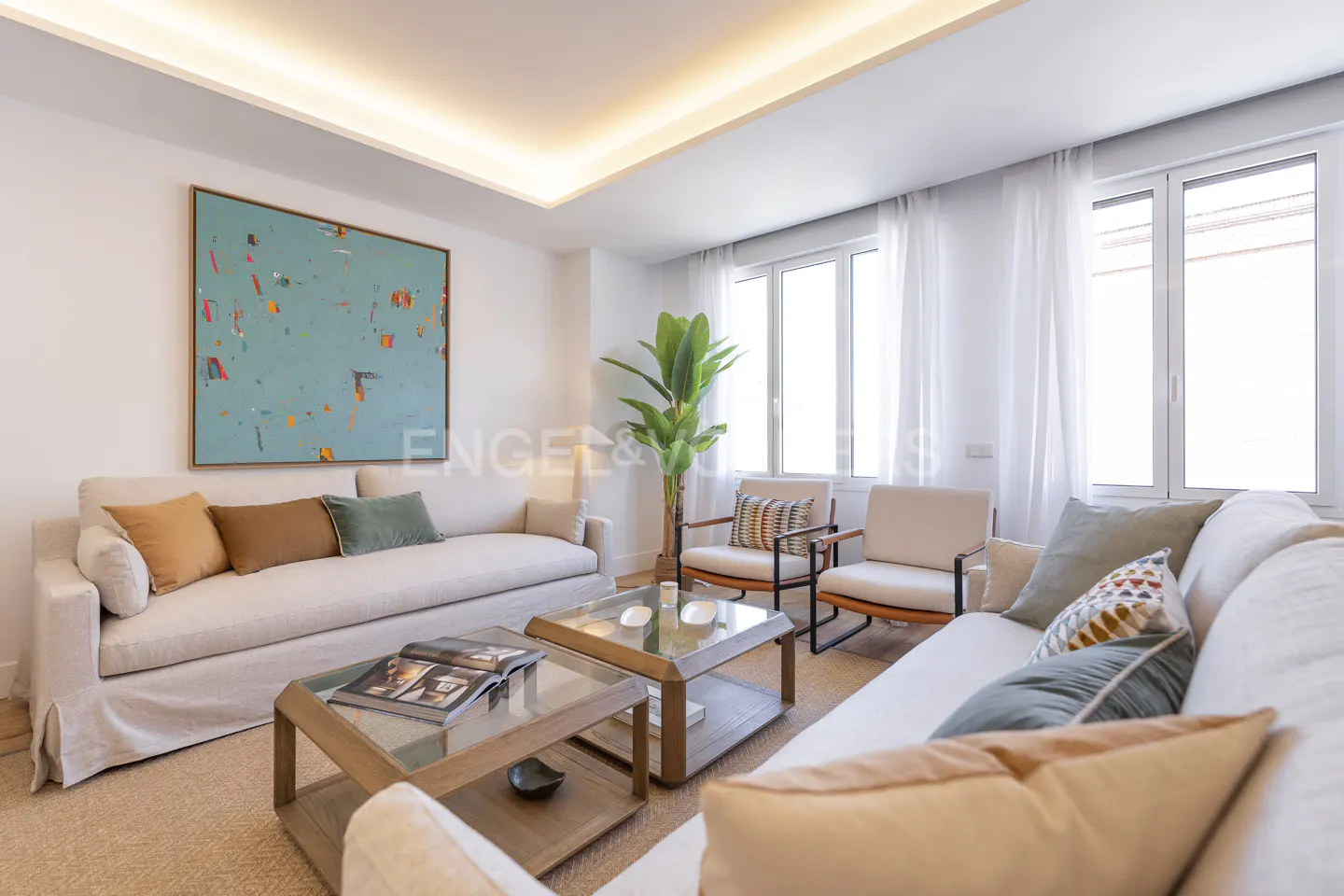 Bright living room with two sofas, two chairs, and two glass-topped coffee tables. A large blue abstract painting hangs on the wall.