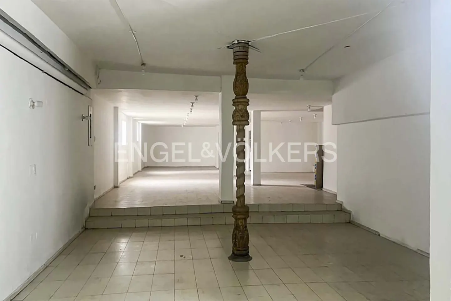 Empty white commercial space with tiled floor, steps, and decorative wooden support column. Engel & Volkers logo in background.