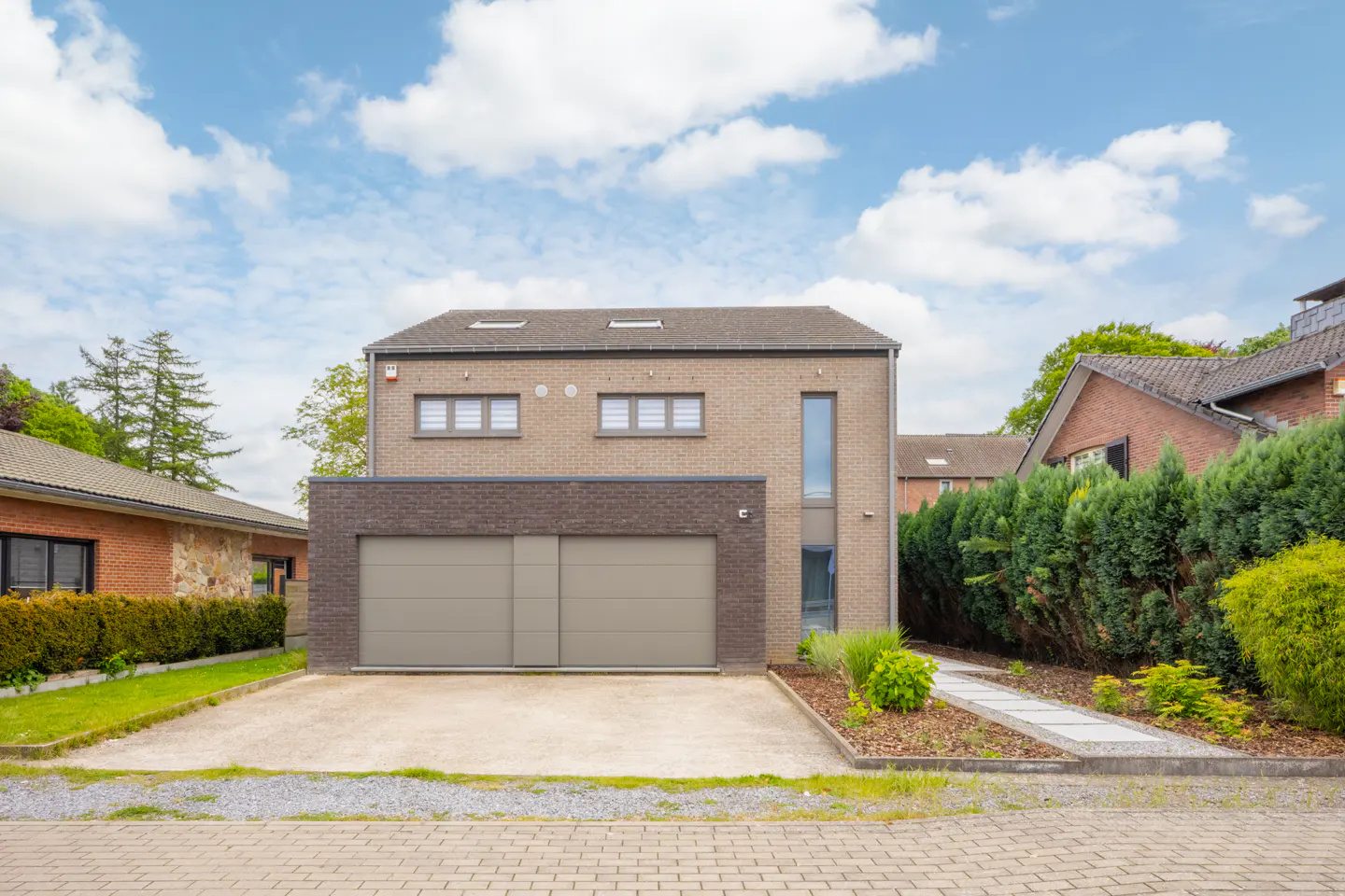 Tan brick two-story house with a double garage and a concrete driveway under a blue sky with white clouds.