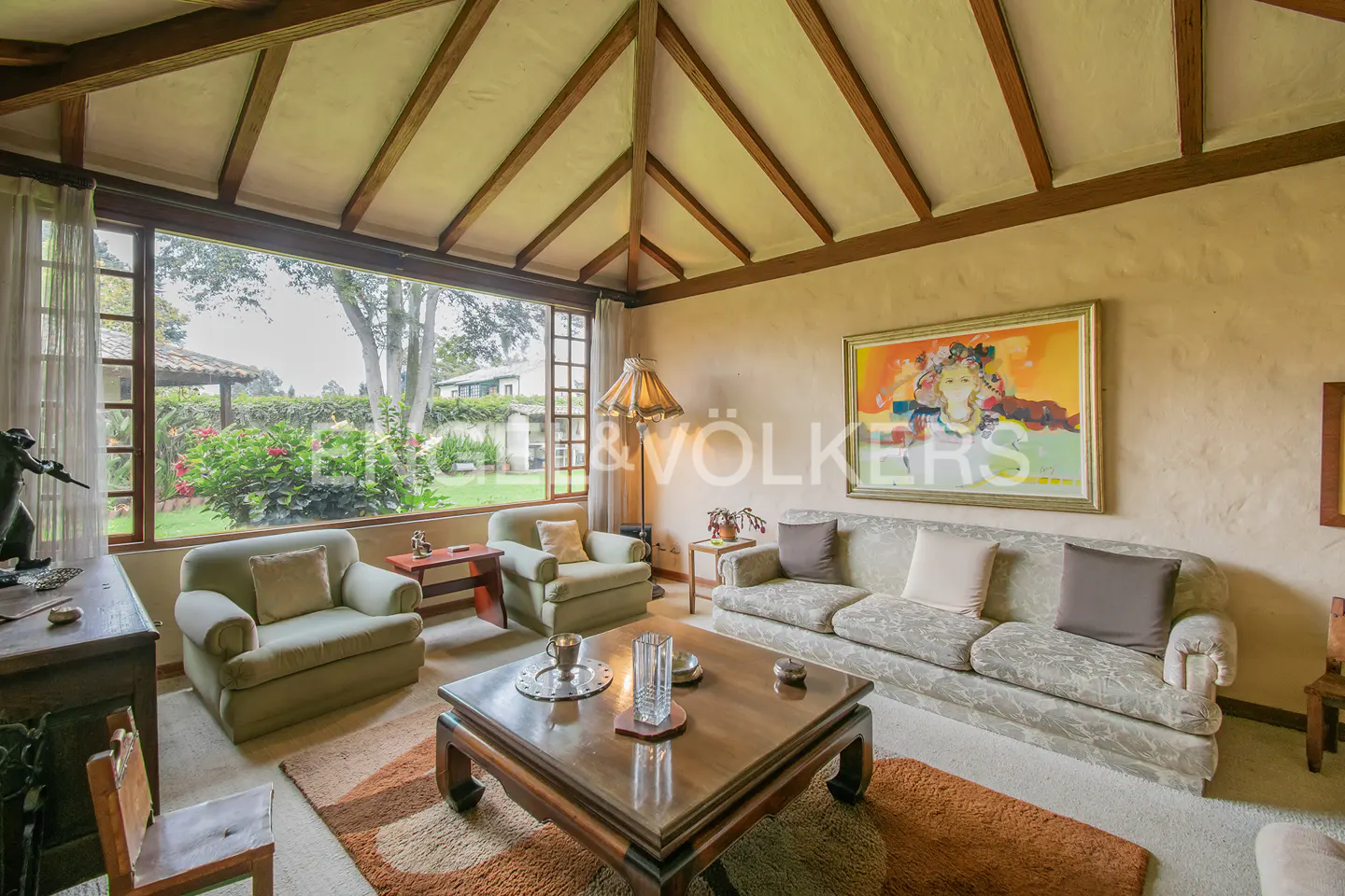 Living room with vaulted wood beam ceiling, large window overlooking garden, sofa, armchairs, and coffee table.