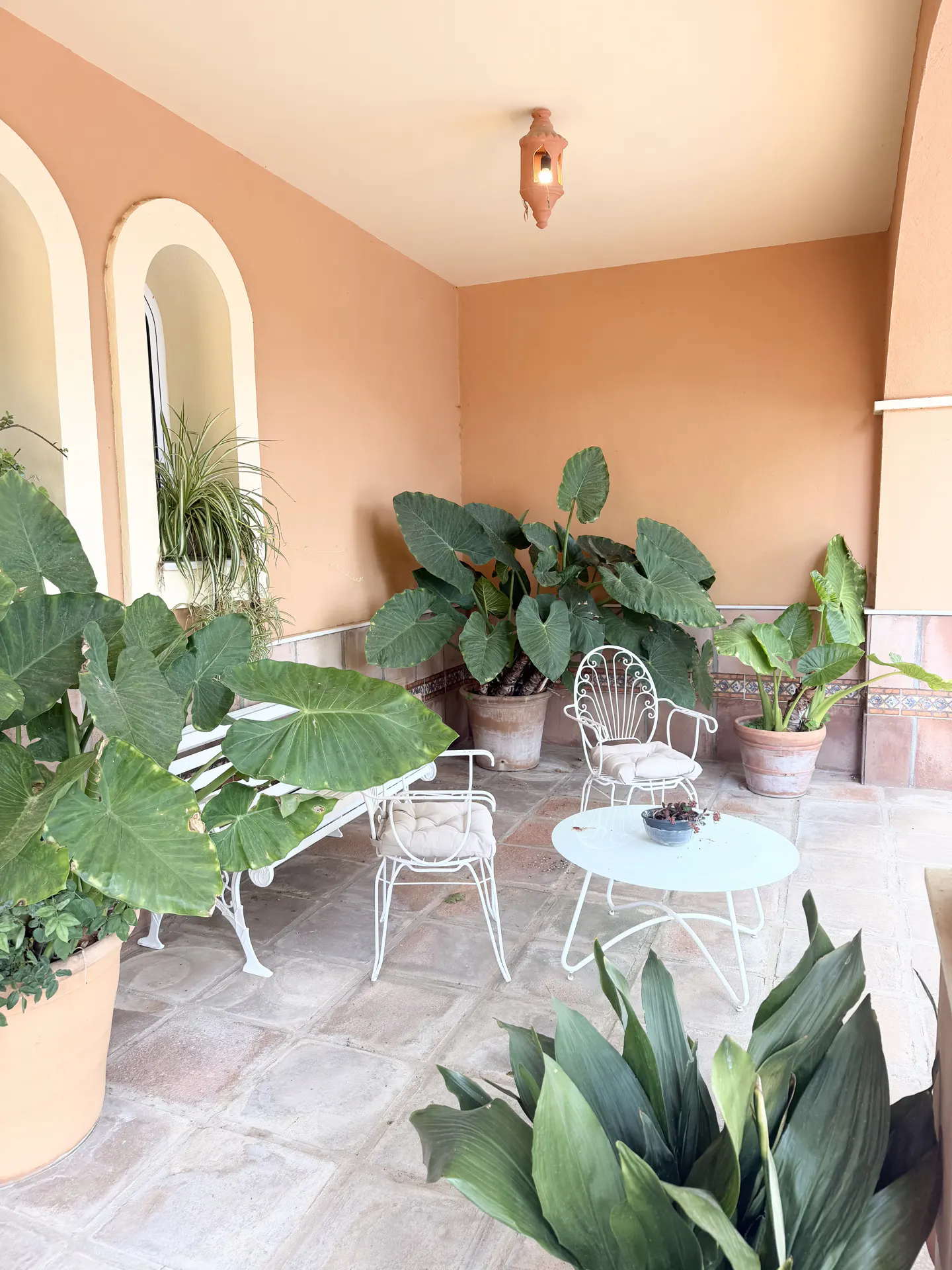Covered patio with terracotta walls, tiled floor, and white wrought iron furniture. Large potted plants add greenery to the space.