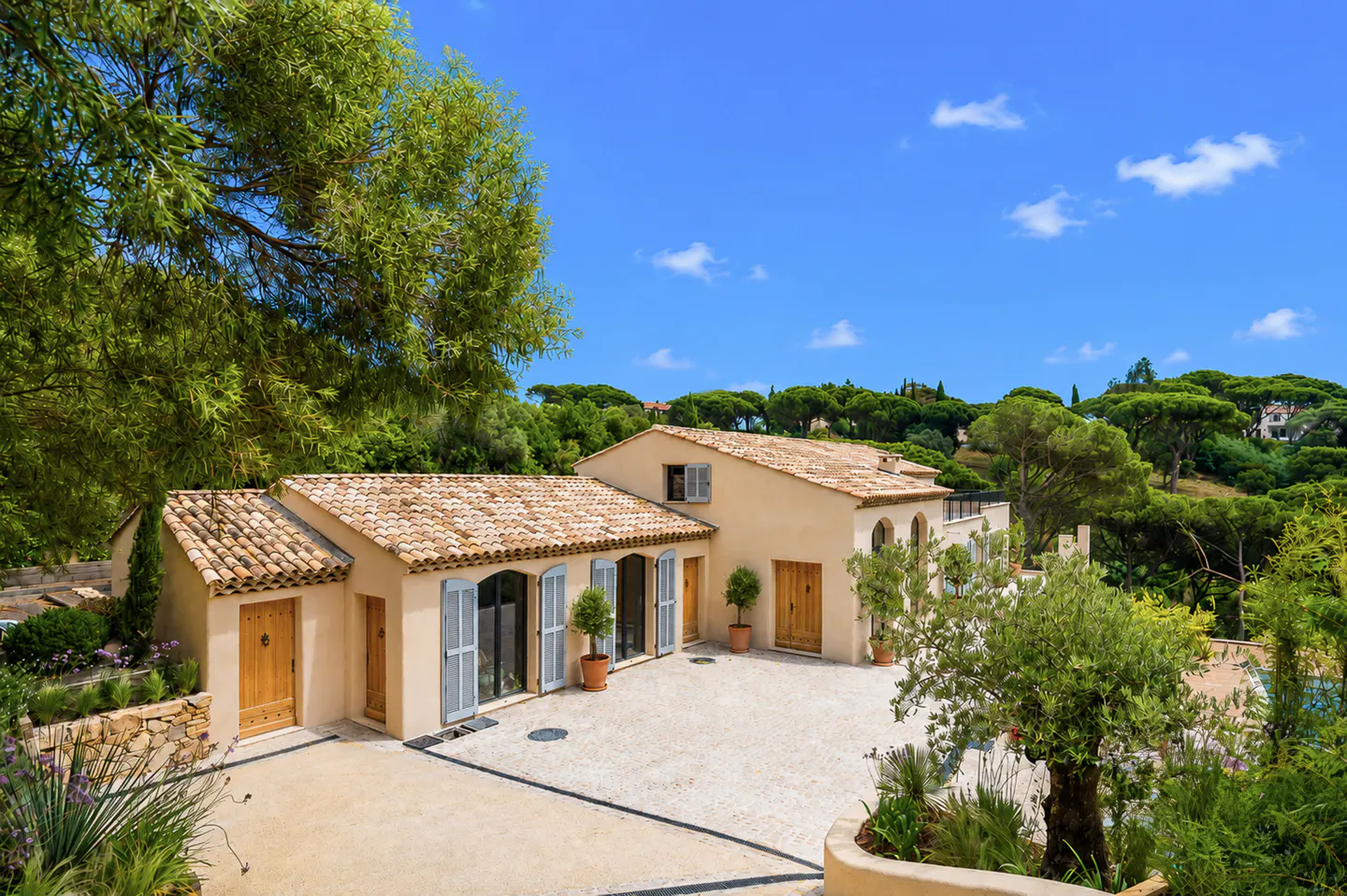 Exterior view of a beige house with a terracotta tile roof, blue shutters, and a gravel courtyard under a blue sky.