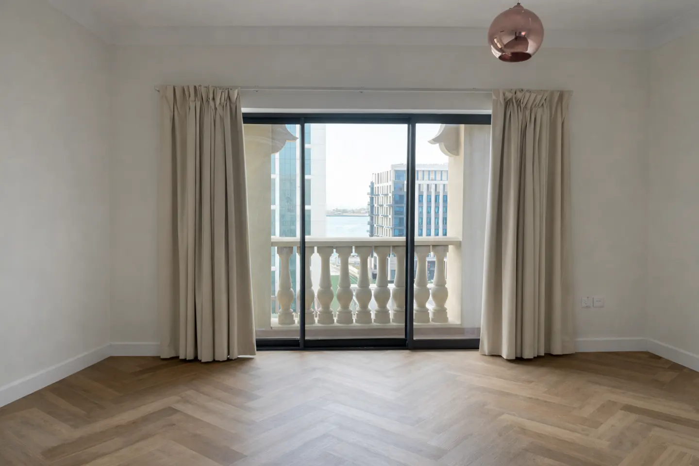 Empty room with herringbone wood floors, white walls, and balcony with city view. Beige curtains frame the sliding glass doors. A copper pendant light hangs above.
