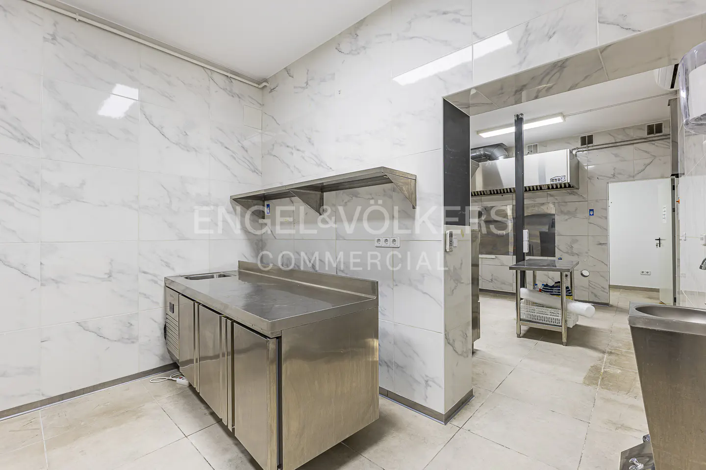 Commercial kitchen with stainless steel prep tables, marble-patterned tiled walls, and a view into a secondary prep area.