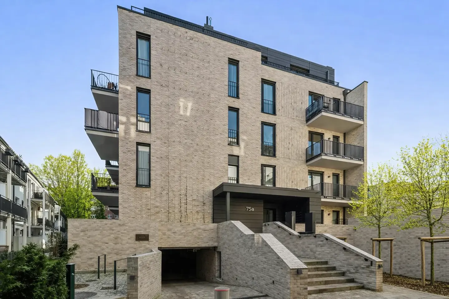 Modern apartment building with light brick facade, black window frames, and balconies. Steps lead to the entrance, with a garage below.